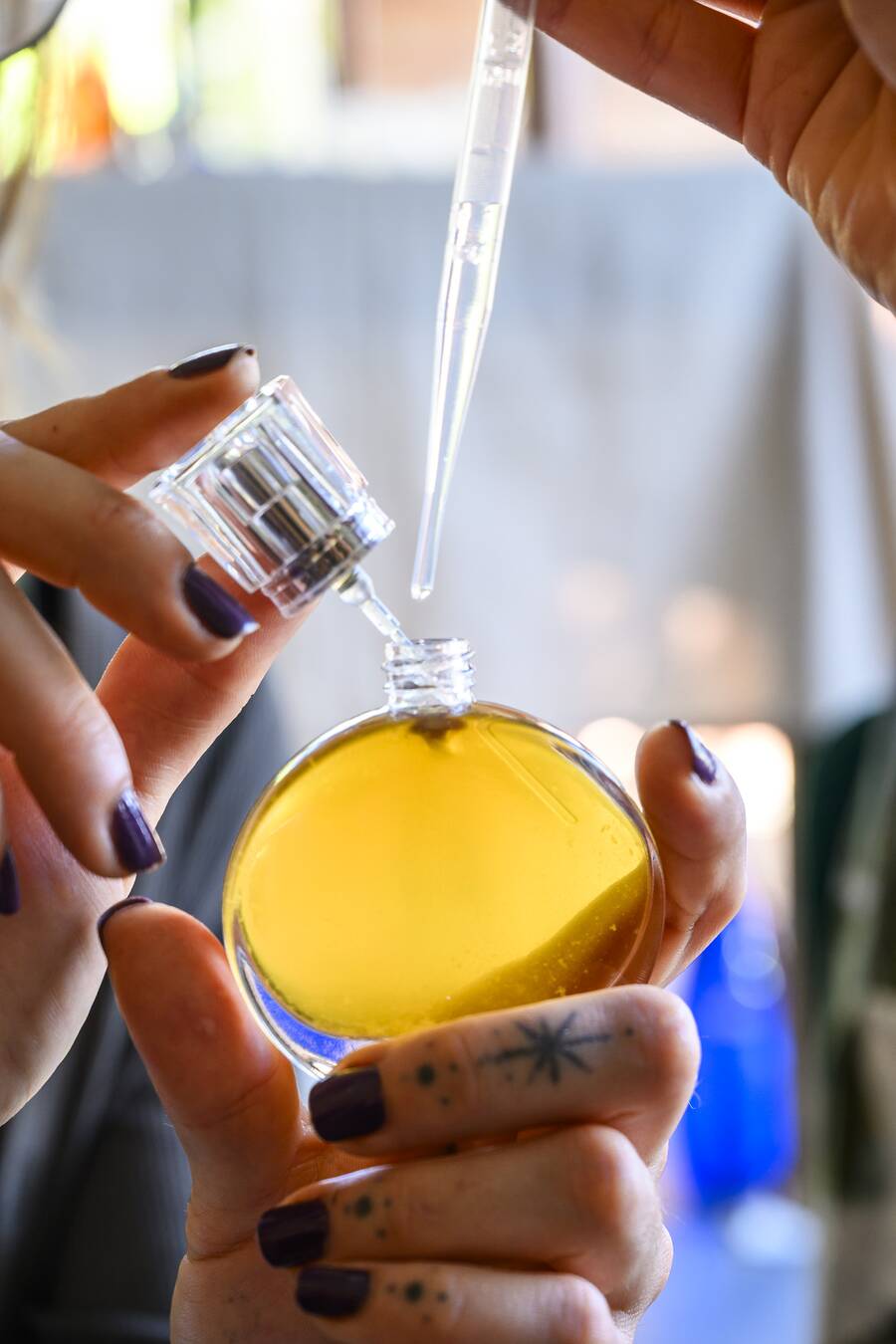 Mãos seguram um frasco redondo com perfume natural dourado, recebendo gotas de uma pipeta